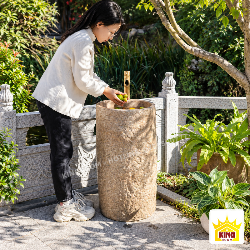 Người phụ nữ cầm trái cây bên chậu rửa đá dáng trụ có vòi màu vàng, đặt cạnh lan can đá trong sân vườn.