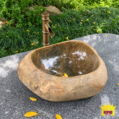 Chậu lavabo đá cuội tự nhiên đặt bàn tông nâu mộc, góc nghiêng trên mặt đá xám có hạt màu, phía sau là cây xanh, lòng chậu có nắp thoát nước màu đồng và lá vàng