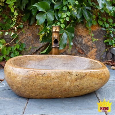 Chậu lavabo đá cuội tự nhiên đặt bàn tông vàng nâu, góc chính diện, thành chậu vàng nâu bo tròn, phía sau là vòi màu đồng và tường đá có cây leo