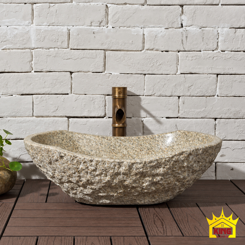 Chậu lavabo đá cuội tự nhiên nguyên khối xám be, góc chính diện, thành đá nhám xám be, đặt trên bàn gỗ nâu trước tường gạch trắng và vòi màu đồng