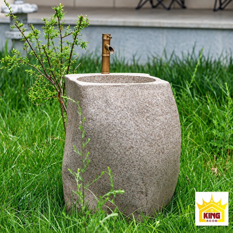 kingroom-chau-lavabo-da-tru-bo-tron-800x800.png Chậu lavabo đá tông xám dáng trụ bo tròn, có vòi màu đồng, đặt trên thảm cỏ cạnh cây xanh, logo KINGROOM ở góc dưới bên phải.