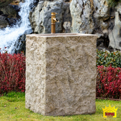 Chậu lavabo khối đứng chữ nhật chụp gần chính diện, có vòi màu đồng, đặt trên thảm cỏ cạnh bụi cây đỏ và tiểu cảnh thác nước