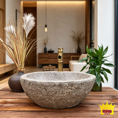 Chậu lavabo xi măng composite giả đá cuội tròn Ø47 × 16 cm chụp chính diện trên bàn gỗ trong nhà, cạnh bình cỏ khô và cây xanh, có vòi màu đồng phía sau