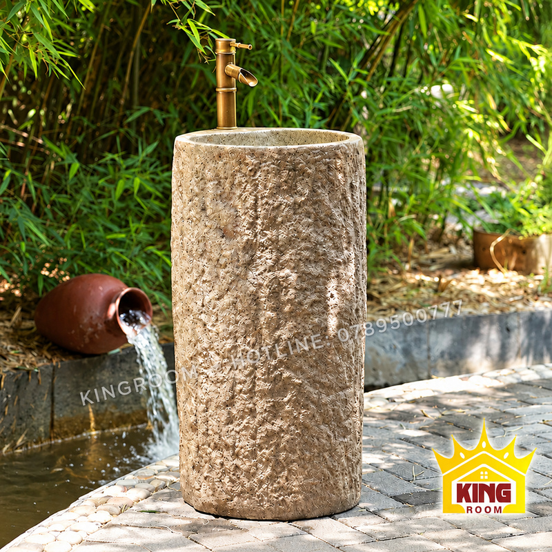 Chậu rửa đá hình trụ cao, bề mặt thô màu be và vòi màu đồng, đặt cạnh dòng nước chảy từ chum trong vườn tre.